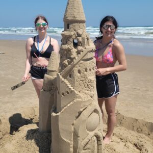 two Teenage girls stand behind a giant 6ft sandcastle on south padre island beach built using Andy Hancock's Verified system of building sandcastles using applied science. They are holding some professional tools supplied during the lesson.