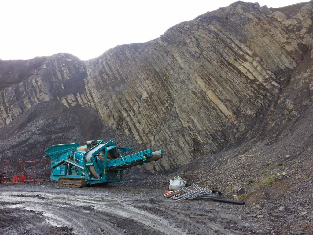 picture showing a large rock crushing machine beneath a 200ft rock face containing unique geological formations in Wales, UK